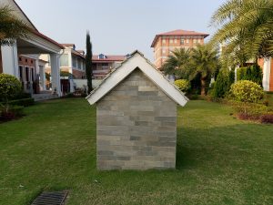 A small, stone-clad house with a peaked roof situated in a well-maintained green lawn. In the background, there are larger buildings and several palm trees. The scene is bright and clear, suggesting a peaceful outdoor environment.