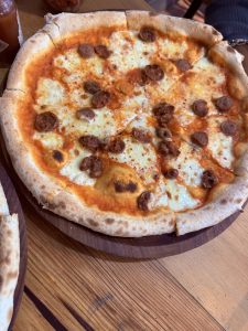A close-up view of a pizza on a wooden plate, featuring a golden-brown crust topped with melted cheese and small pieces of sausage. 