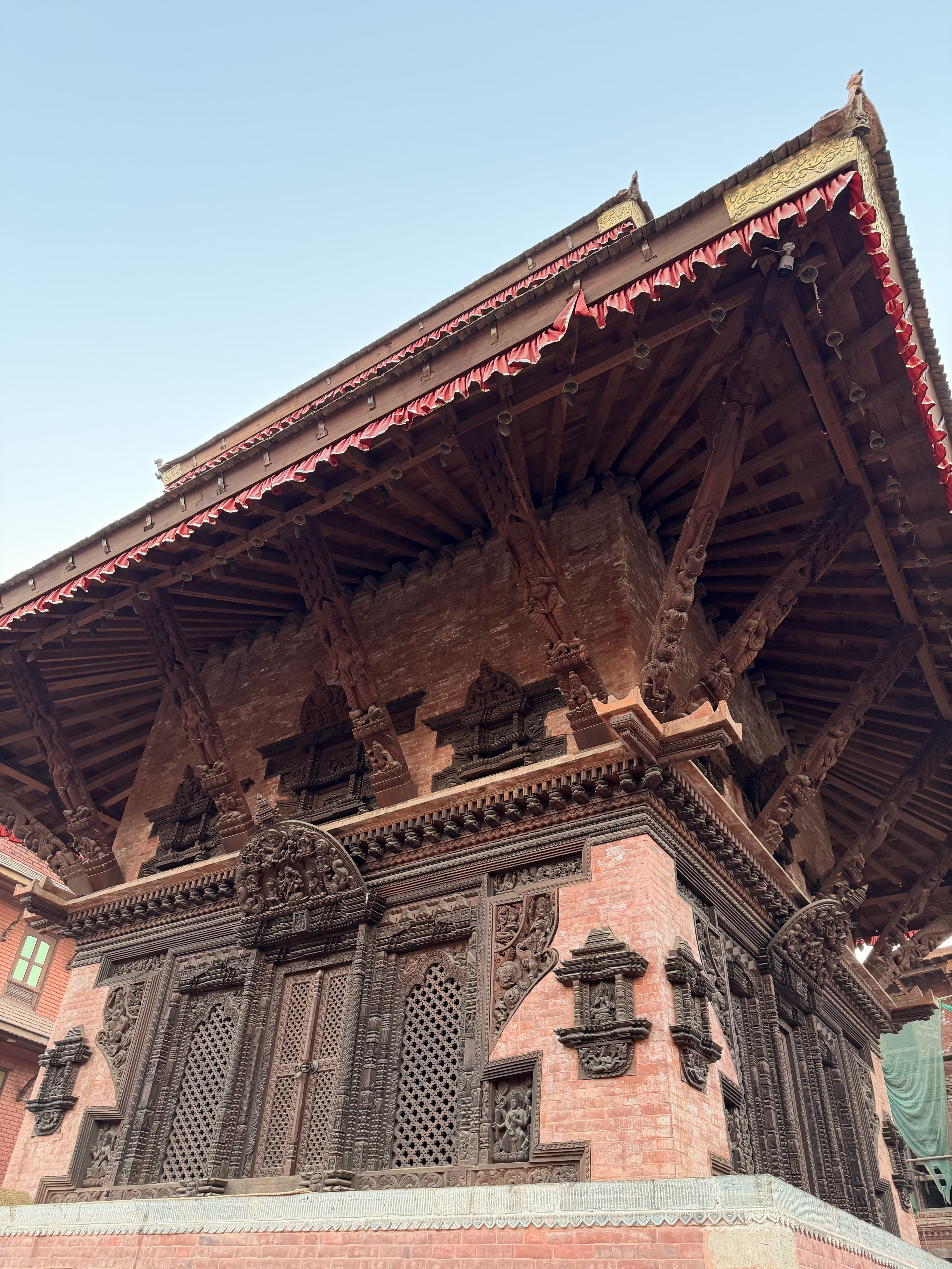 A detailed view of a traditional architectural structure featuring intricate wood carvings and brickwork. The building has an elaborate wooden roof with red decorative elements and a series of carved panels on the walls showcasing artistic designs.