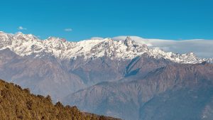 A panoramic view of majestic snow-capped mountains under a clear blue sky, with rocky slopes in the foreground and patches of greenery.