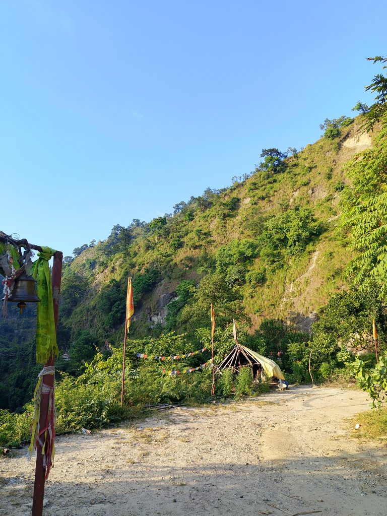 A scenic view of a rural landscape featuring a dirt path leading up to a small, makeshift hut surrounded by greenery. Brightly colored flags and a bell are visible in the foreground, while steep hills covered in lush vegetation rise in the background under a clear blue sky