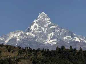 A majestic snow-capped mountain rises prominently against a clear blue sky, surrounded by rugged peaks and green hills.