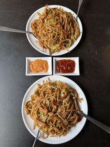 Two plates of chowmein noodles topped with savory sauce, presented side by side on a wooden table.