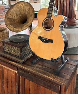 A wooden acoustic guitar is displayed on a wooden table alongside a vintage gold-colored gramophone and a decorative box. 