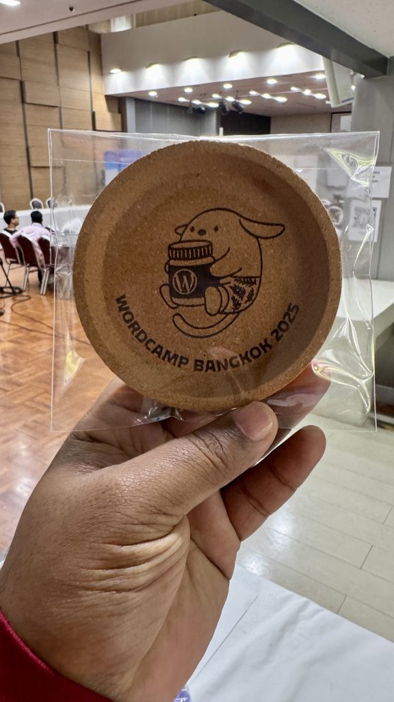 A person holding a round cork coaster featuring a cute dog illustration and the text “WordCamp Bangkok 2025” in a clear plastic wrapper.
