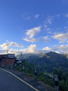 A scenic view of a mountainous landscape under a blue sky with scattered clouds. In the foreground, there is a road with a curved sign indicating a turn ahead