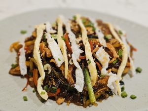 A plate of glossy Korean stir-fried noodles mixed with vegetables and meat, topped with thin egg strips, sesame seeds, and spring onions. The dish looks rich, colourful, and freshly served. Captured from Kozhikode, Kerala. 