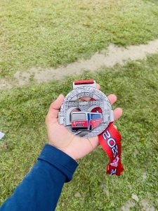 A hand wearing a blue sleeve holds a metallic finisher medal with a red and white ribbon. The medal features a stadium design and a sports bus. The background shows grassy terrain.