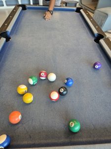 A close-up of a billiards table with colorful pool balls on blue felt and a hand reaching in during play.
