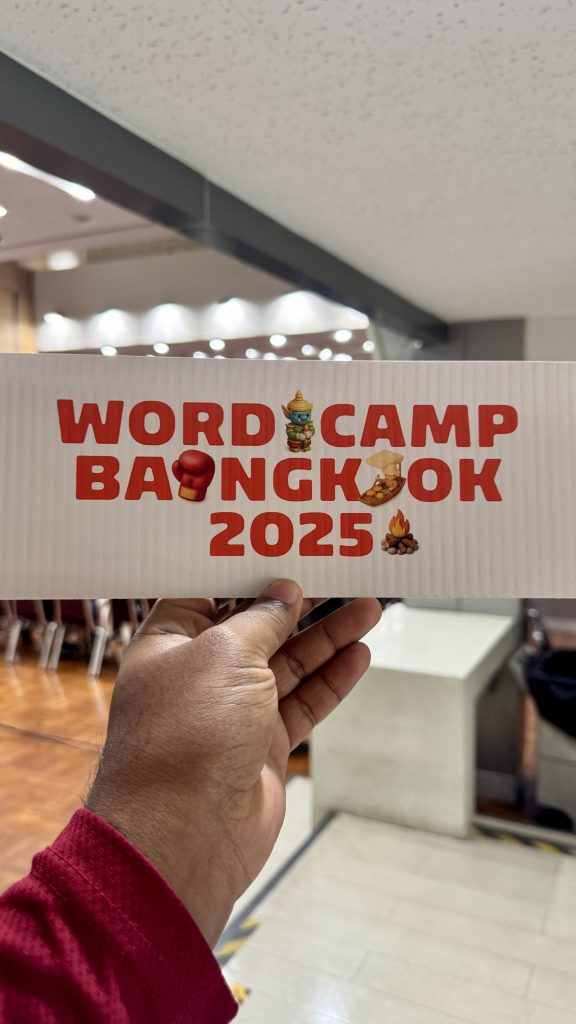 A person holding a rectangular white card with “WordCamp Bangkok 2025” printed in red letters, decorated with Thai-themed icons like a boxing glove and a floating market boat.
