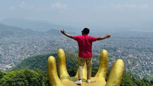 A person standing with arms outstretched on a large golden hand sculpture, with a city and mountains of Nepal in the background.