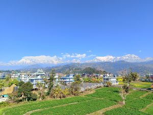 
A panoramic view of a town featuring colorful buildings and greenery in the foreground, set against a backdrop of majestic snow-capped mountains under a clear blue sky. 