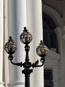 A decorative street lamp with three glass globes sits against a backdrop of white columns and a building facade.
