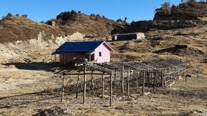 A pink house with a blue roof sits in a dry, hilly landscape. Nearby, a partially constructed wooden structure is visible, along with some scattered twigs and brush.
