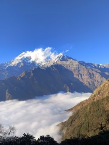 A majestic mountain landscape features snow-capped peaks under a clear blue sky. Below, a thick blanket of clouds covers the valley, creating a dramatic contrast with the rugged terrain. 