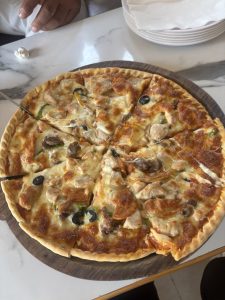 
A freshly baked pizza on a wooden serving board, topped with pieces of chicken, black olives, green bell peppers, and mushrooms, with melted cheese stretching from a slice that has been partially cut.