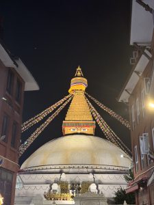 
A large, illuminated stupa with colorful prayer flags hanging from its top, set against a dark night sky. 
