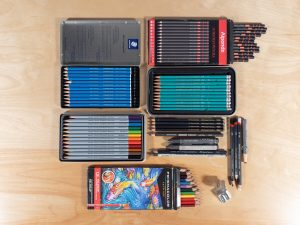 A top view photograph of several types of drawing pencils ranging from graphite, woodless pencils, matt pencils, colored pencils, and watercolor pencils, along with a pencil sharpener.