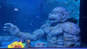 A massive, moss-covered stone statue of a mythical giant submerged in an aquarium surrounded by swimming fish.