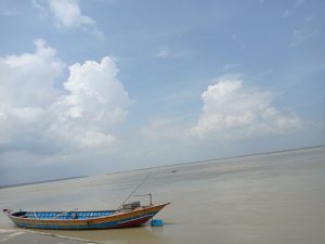 A traditional wooden boat resting along the Padma River under a bright blue sky with scattered clouds, capturing the calm and vast river landscape of Bangladesh.