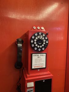 A vintage red rotary payphone mounted on an orange wall. It has a black handset, numbered dialing wheel, and a sign indicating call costs. Retro and nostalgic tone.