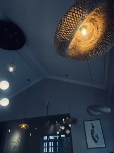 The image features a ceiling with various pendant lights in different styles. A large woven design light hangs prominently, casting a warm glow. Smaller round lights, some with a glass covering, are arranged nearby.