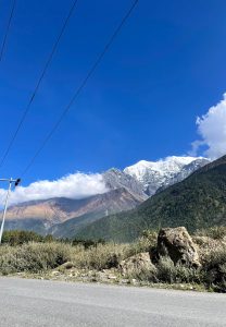 A scenic landscape featuring majestic snow-capped mountains under a clear blue sky with Lush greenery and rocky formations are visible in the foreground, with power lines running diagonally across the image.