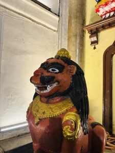 A stylized statute of a lion. It's reddish-brown with a black mane that flows like straight hair. It has black markings on its face, and it's teeth are bared, though no menacingly. It's wearing a gold necklace and arm jewelry. The setting appears to be a temple.