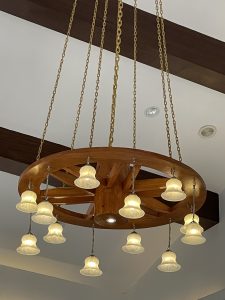 A circular wooden chandelier resembling a wagon wheel, with twelve bell-shaped frosted glass lights, hanging from gold chains amid white ceiling and dark wooden beams.