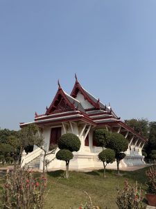 A traditional Thai-style building with intricate red and gold architectural details, surrounded by manicured shrubs and trees.