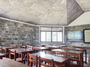 A classroom featuring stone walls and a uniquely designed ceiling. The room is equipped with wooden desks and chairs arranged in rows.