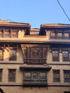 A close-up view of a traditional brick building featuring intricate wooden window structures and ornate carvings. The upper part of the building showcases multiple gabled roofs, while wooden latticed windows are visible throughout. The scene is set against a clear blue sky, highlighting the architectural details and craftsmanship of the structure.
