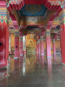 
An ornate interior of a temple featuring vibrant red and gold columns, intricately decorated ceilings, and a large Buddha statue at the far end.