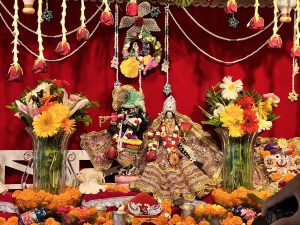 A vibrant altar with two dressed deities, surrounded by flowers, marigold garlands, offerings, and festive decorations against a red backdrop.