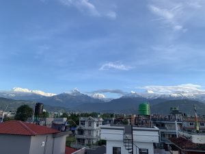 A panoramic view of snow-capped mountains under a clear blue sky, with a mix of residential buildings in the foreground. 