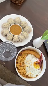 A wooden table is set with two dishes. One circular white plate features a ring of steamed momo (Nepali-style dumplings) surrounding a small bowl of sauce. Next to it is a bowl containing yellow noodles topped with a fried egg and a piece of sausage. A glass of water is positioned nearby, and there is a small white vase with a green plant. The setting is casual and inviting.