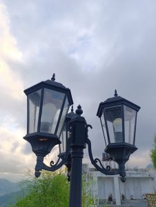 A black, vintage-style lamp post with three lanterns stands against a backdrop of cloudy skies. 
