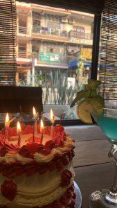 A beautifully decorated birthday cake with red frosting and cherries on top sits in the foreground, featuring several lit candles. 
