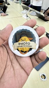 A person's hand holding a round white button badge featuring a cute yellow dog character and the text "PHOTO CAPTURE THE MOMENT."