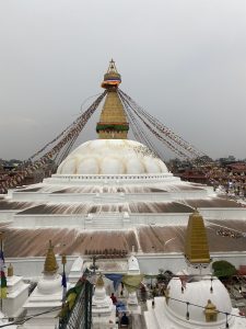 A large white stupa adorned with colorful prayer flags, set against a cloudy sky. The stupa features a golden tower with large eyes painted on its facade, representing a sense of watchfulness.