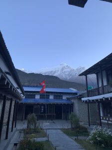 A peaceful mountain courtyard with traditional buildings, snow-capped peaks, and Nepali flags under a clear blue sky.