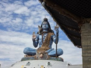 A large, colorful statue of Shiva with blue skin, a serpent necklace, crescent moon in his hair, seated on a tiger-shaped throne, holding a trident, with a cloudy sky and traditional building in the background.