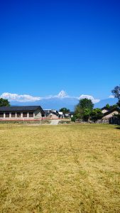 Vertically oriented, the foreground is a broad grass lawn. In the middle are some buildings and trees. In the distance are giant mountains covered in snow.  The top of the image is a wide clear sky.