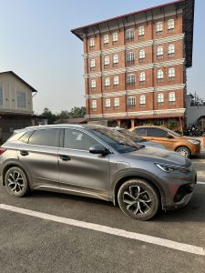A grey SUV is parked in the foreground on a street, displaying some dirt on its side. In the background, there is a multi-story brick building with large windows and a red roof
