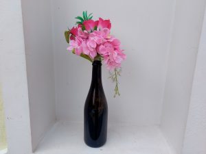 
A dark glass bottle is placed on a white ledge, with a cluster of artificial pink flowers and green leaves arranged in the neck of the bottle