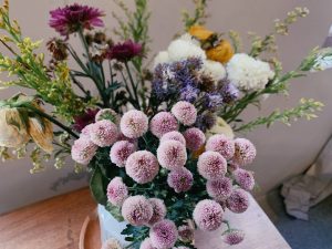 Mixed bouquet of small pink pom-pom chrysanthemums, white and yellow blooms, and purple flowers arranged in a white vase on a wooden table.
