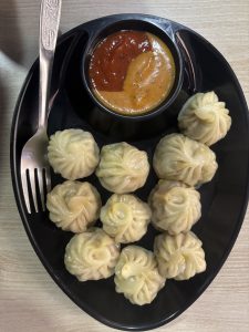 A black plate containing a serving of steamed dumplings arranged neatly, with a small bowl of two types of dipping sauce&mdash;one reddish-brown and the other light brown&mdash;placed in the center.