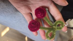 A hand holding a colorful craft item made from pipe cleaners, featuring two red rose-shaped blossoms attached to a green spiral stem.
