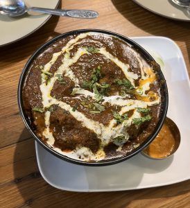 

Bowl of dark brown Mutton curry with yogurt swirls and chopped green herbs.