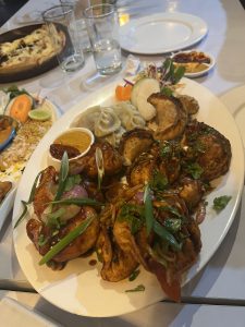 A large platter with assorted dishes, including grilled chicken, dumplings, rice, vegetables, and sauce, set on a dining table with drinks and pizza in the background.